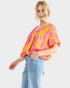 A woman modeling a bright yellow and pink patterned blouse with short flutter sleeves, paired with light blue jeans, against a light gray studio background.