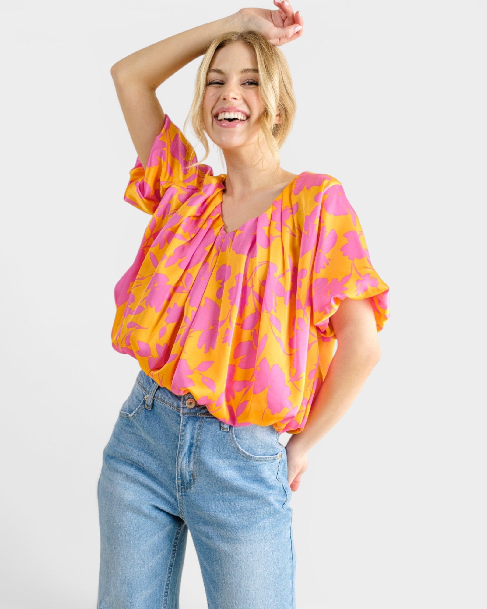 A woman modeling a bright pink and orange patterned short-sleeve blouse with a gathered, draped neckline, paired with light blue jeans against a light gray studio background.