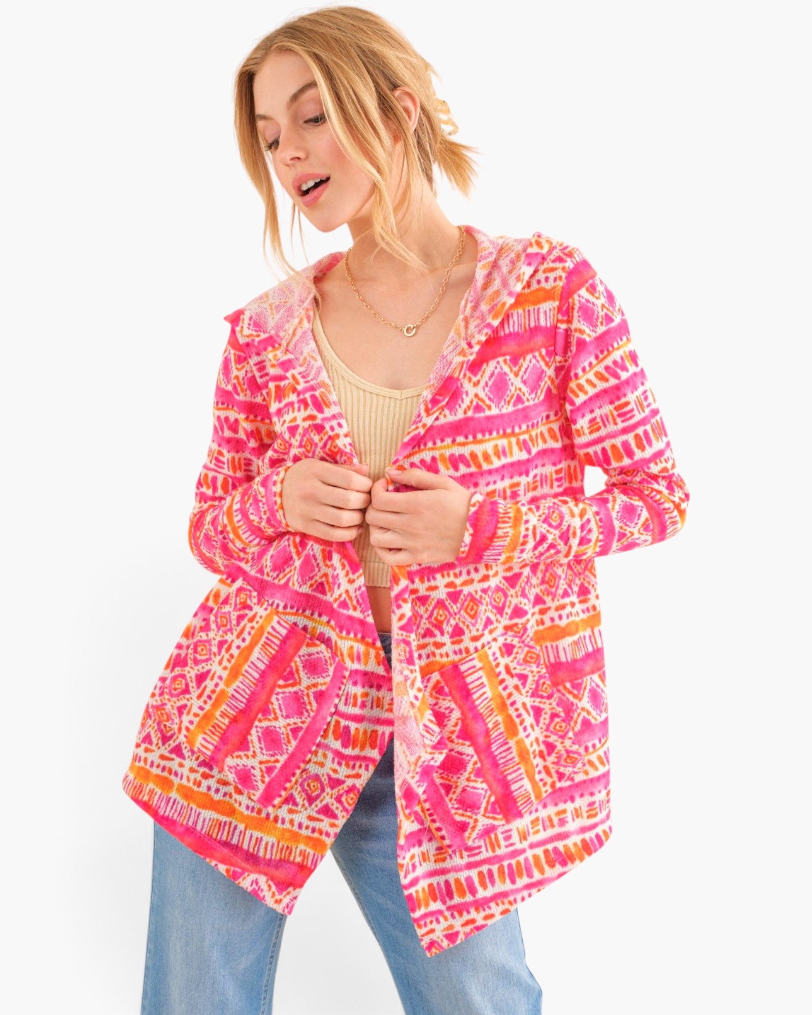 A woman wearing a bright pink, orange, and white patterned cardigan over a beige top and blue jeans, photographed against a plain white studio background.