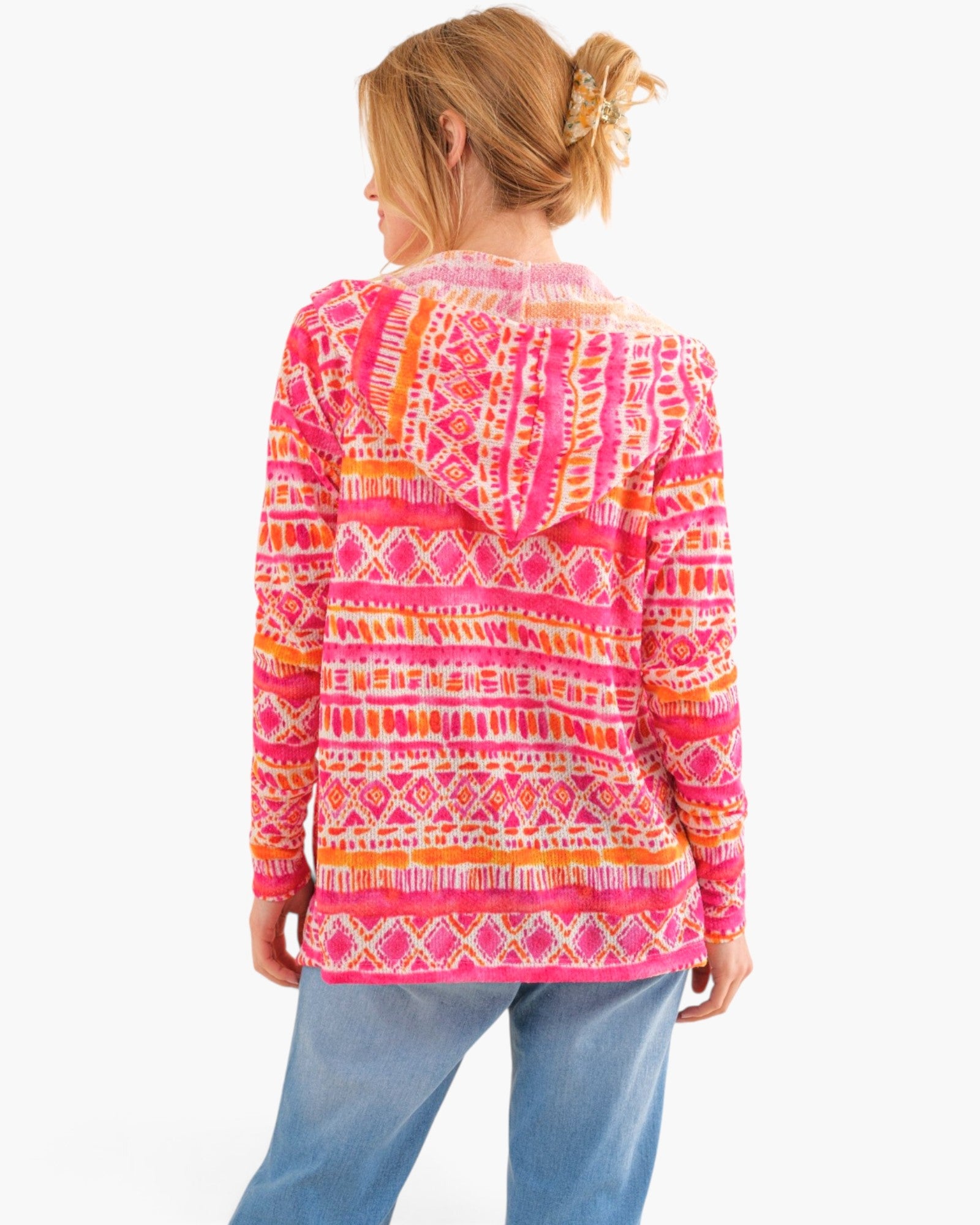 A woman shown from the back wearing a bright pink hooded long-sleeve top with an orange and white geometric pattern, paired with blue jeans, against a plain white background.