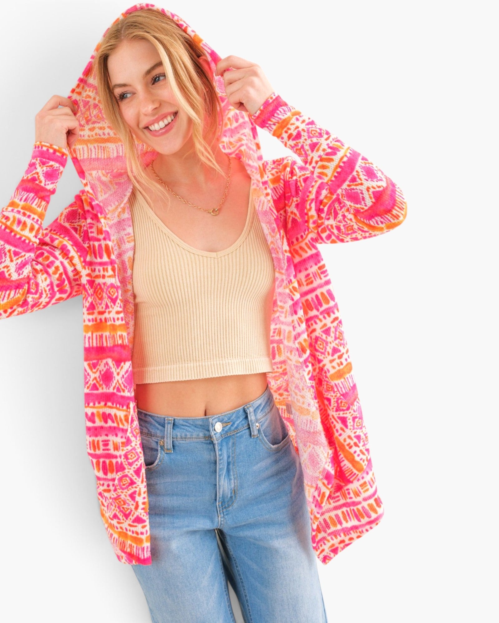 A woman wearing a bright pink, orange, and yellow patterned hooded cardigan over a beige ribbed crop top and blue jeans, posing against a light gray studio background.