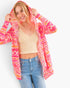 A woman wearing a bright pink, orange, and yellow patterned hooded cardigan over a beige ribbed crop top and blue jeans, posing against a light gray studio background.