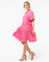 A woman modeling a bright pink short-sleeve tiered dress with a collared neckline and front pockets, paired with beige heeled sandals, against a plain white studio background.