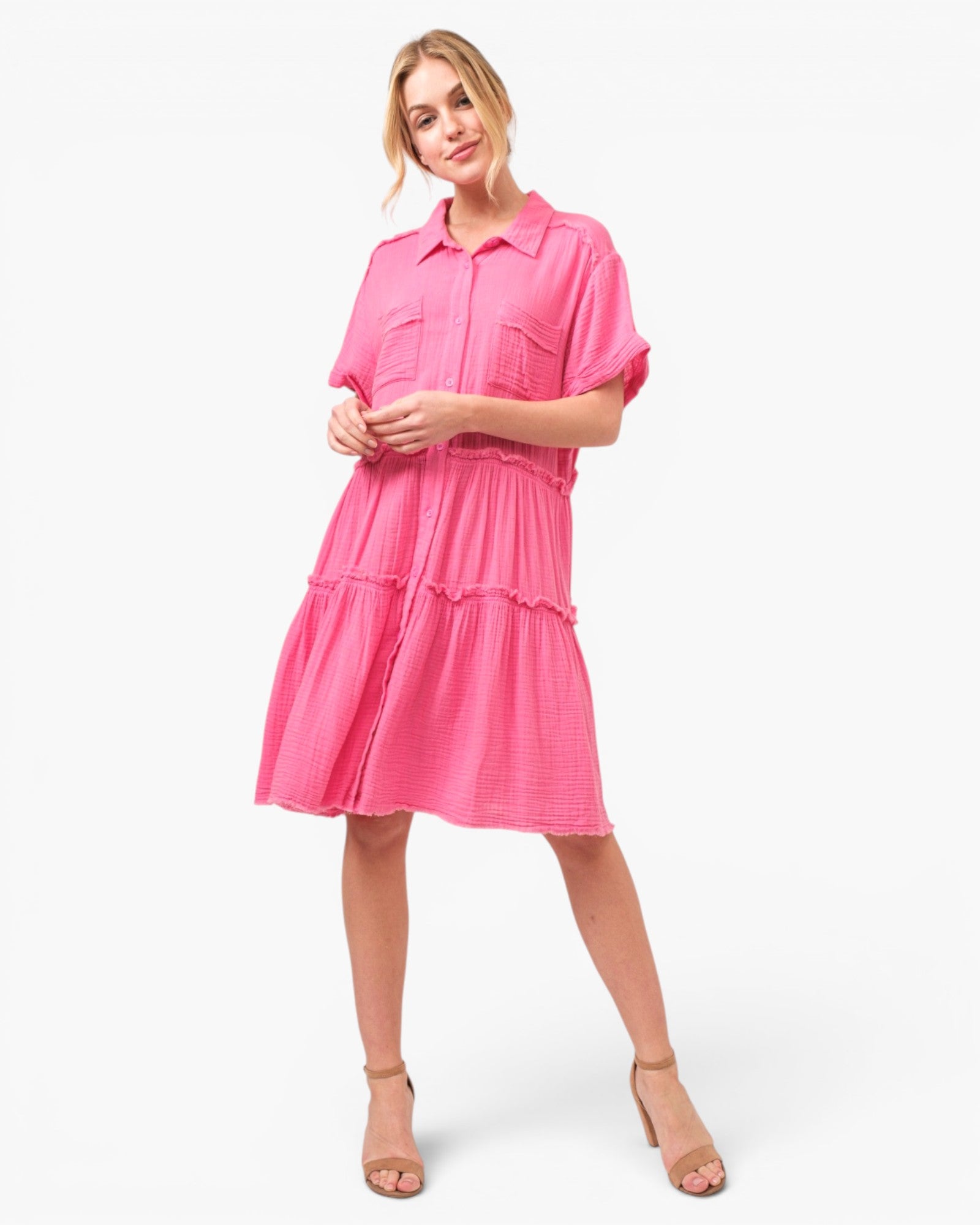 A woman modeling a bright pink tiered shirt dress with short sleeves and a button-up collar, paired with beige high-heeled sandals, against a plain white studio background.