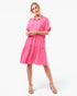 A woman modeling a bright pink tiered shirt dress with short sleeves and a button-up collar, paired with beige high-heeled sandals, against a plain white studio background.