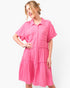 A woman modeling a short-sleeve pink tiered shirt dress with a button-down front and chest pockets, photographed against a plain white studio background.