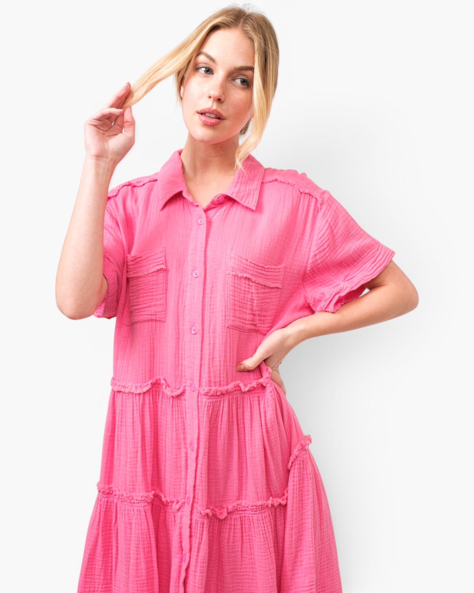 A woman modeling a bright pink short-sleeve button-front dress with a collar, chest pockets, and tiered ruffle details against a plain light background.