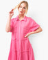 A woman modeling a bright pink short-sleeve button-front dress with a collar, chest pockets, and tiered ruffle details against a plain light background.