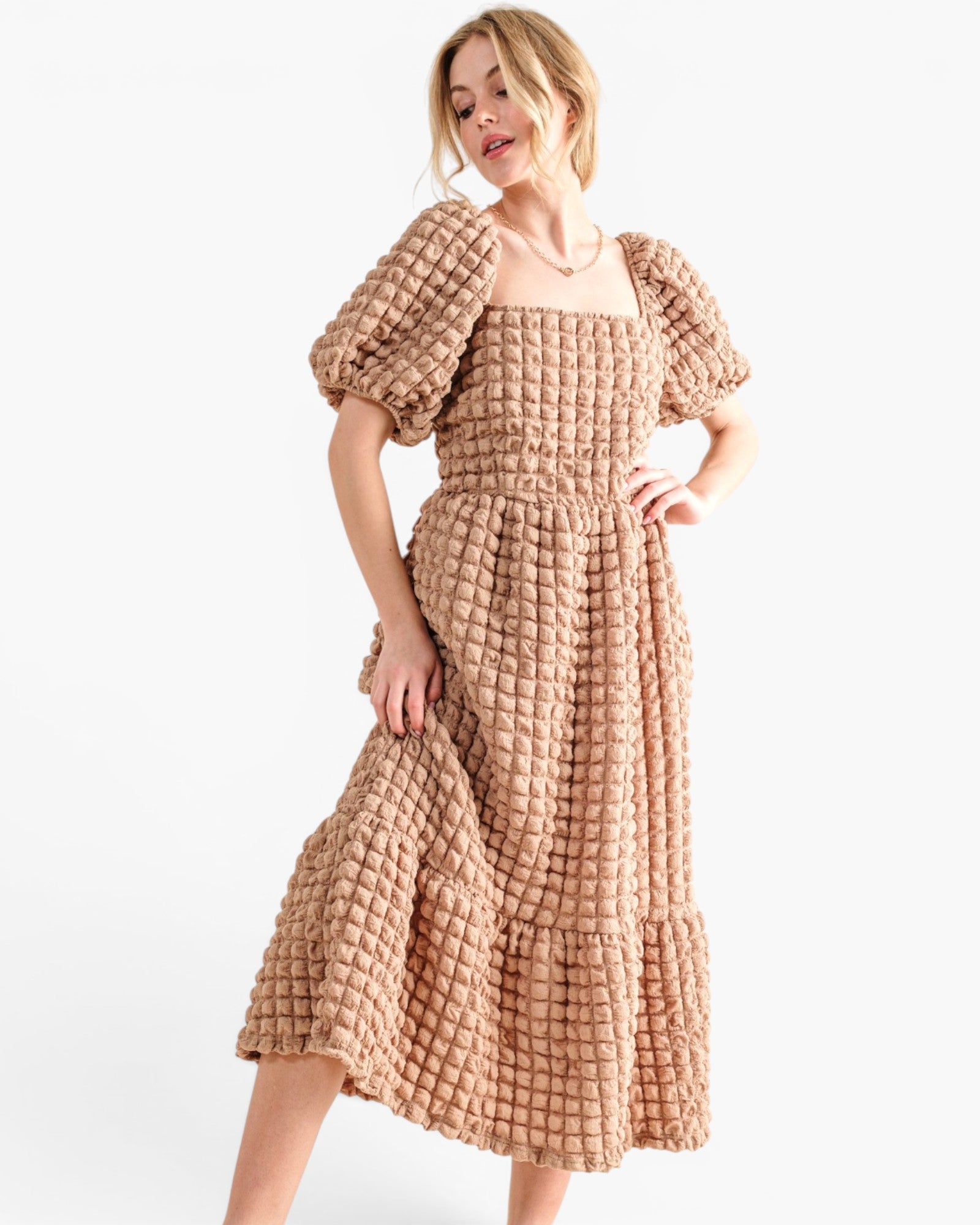 A woman modeling a textured, quilted midi dress in a warm tan/beige color, featuring puff sleeves and a square neckline, set against a plain white studio background.