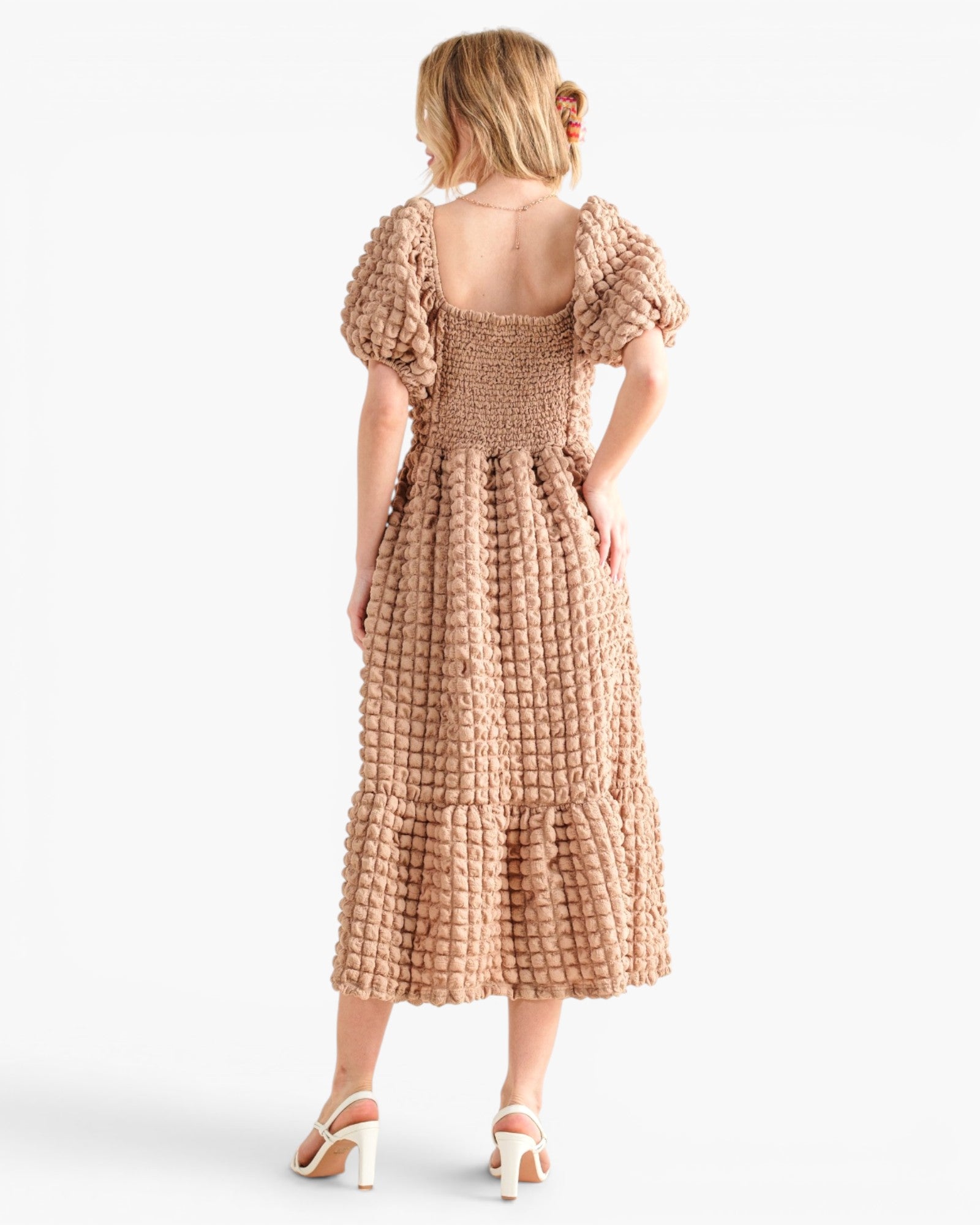 A woman shown from the back wearing a textured, boucle-style midi dress in a warm tan/beige color. The dress features puff sleeves, a smocked bodice, and a tiered skirt.