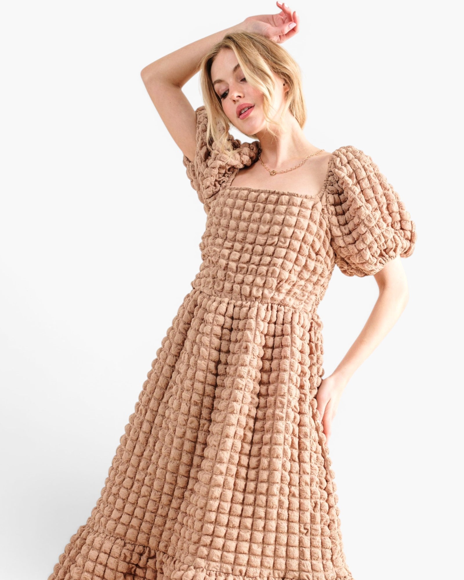 A woman modeling a textured, chunky knit midi dress in a warm beige color, featuring puff sleeves and a square neckline, set against a plain white studio background.