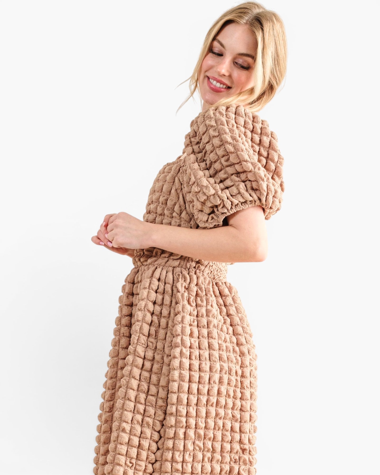 A woman wearing a chunky, quilted short-sleeve dress in a warm beige or light brown color, photographed against a clean white studio background.