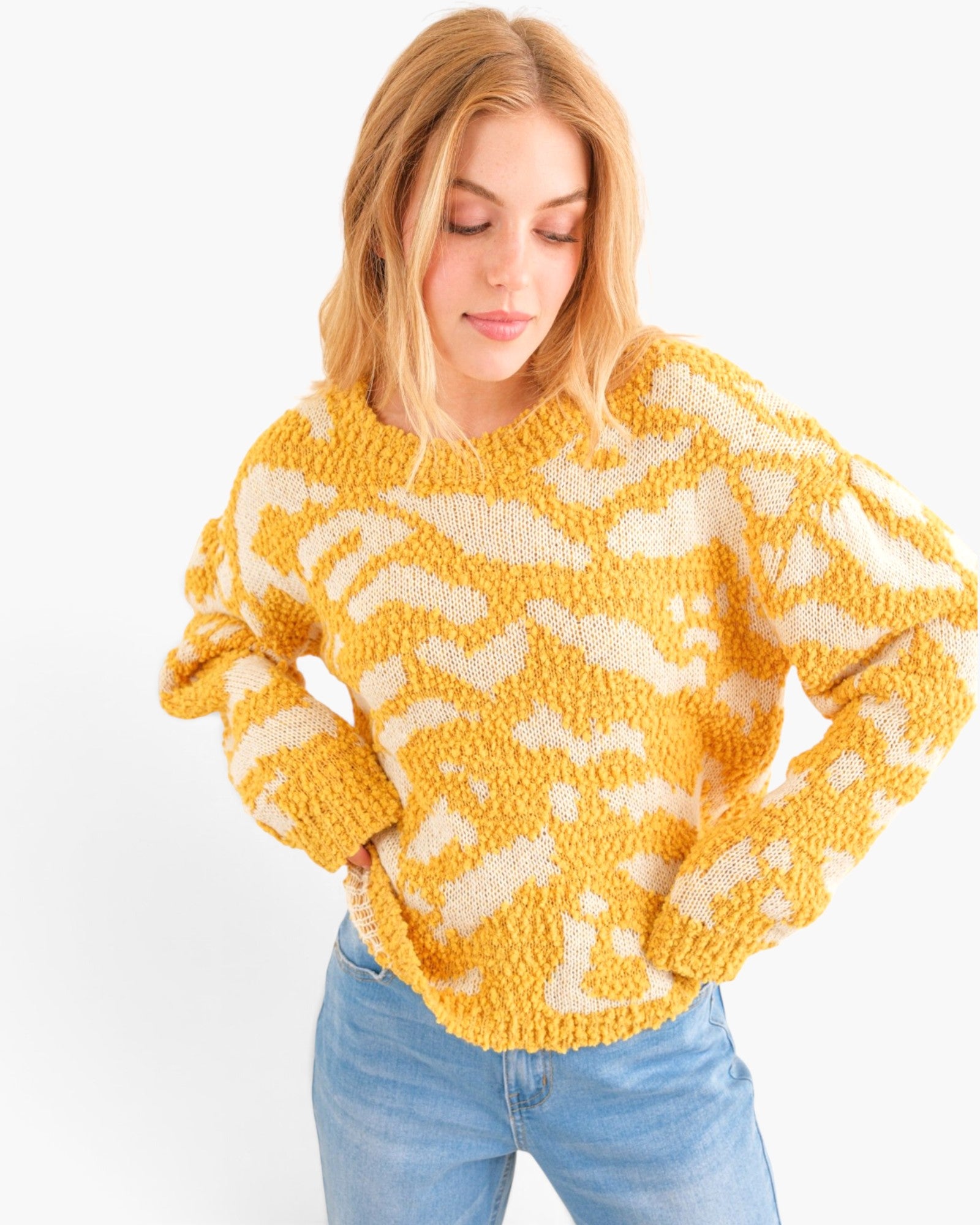 A woman wearing a chunky, textured knit sweater with a yellow and cream abstract pattern, paired with light blue jeans, against a plain white background.