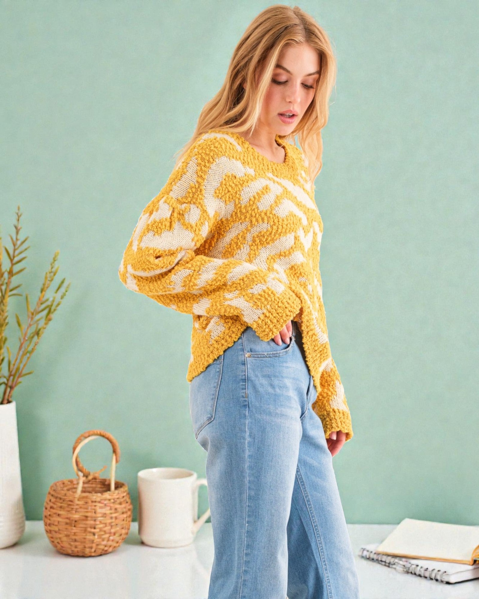 Woman wearing a yellow and white patterned sweater with blue jeans against a light green background.