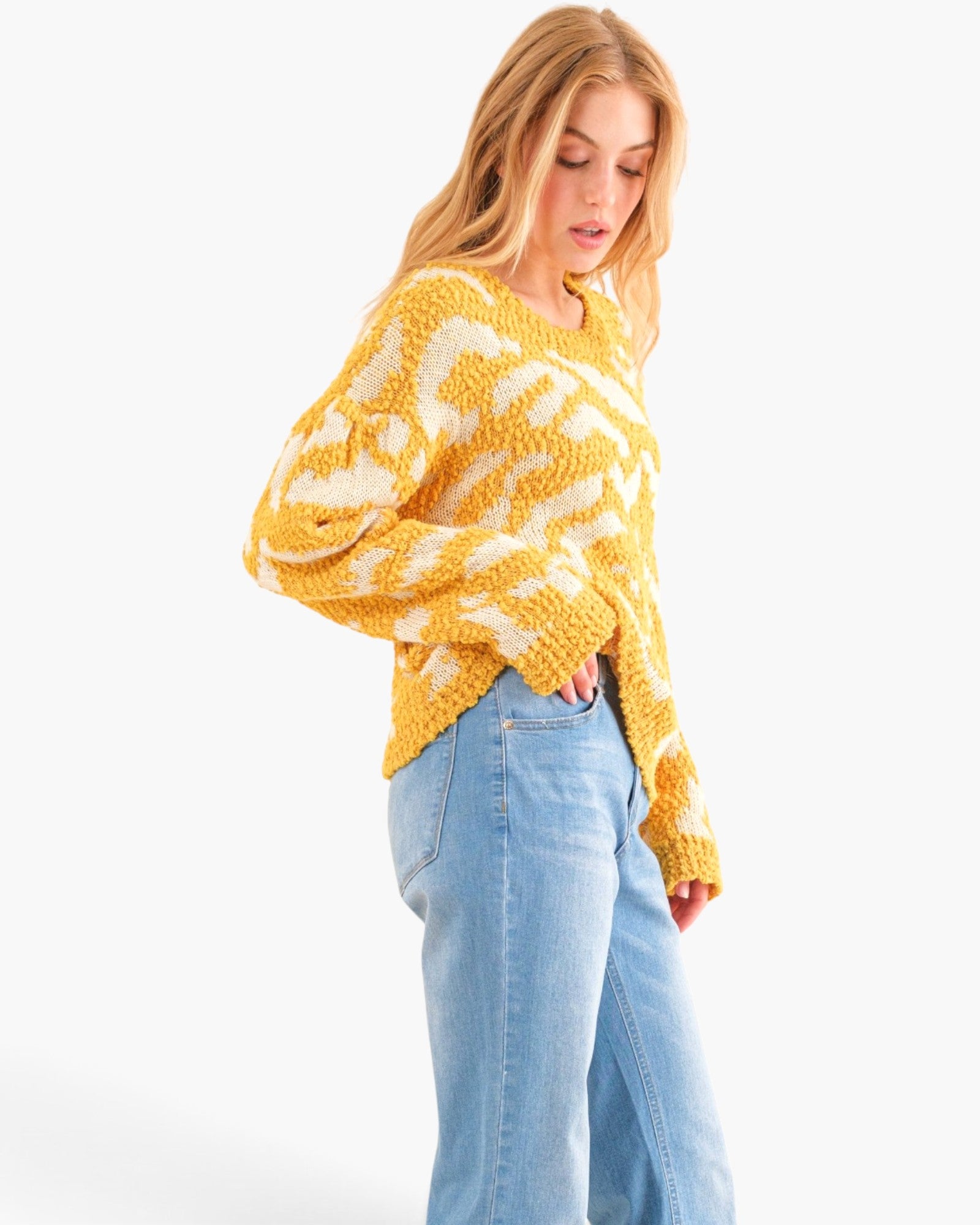 A woman wearing a chunky knit yellow and white patterned sweater with light blue jeans, photographed against a plain white studio background.