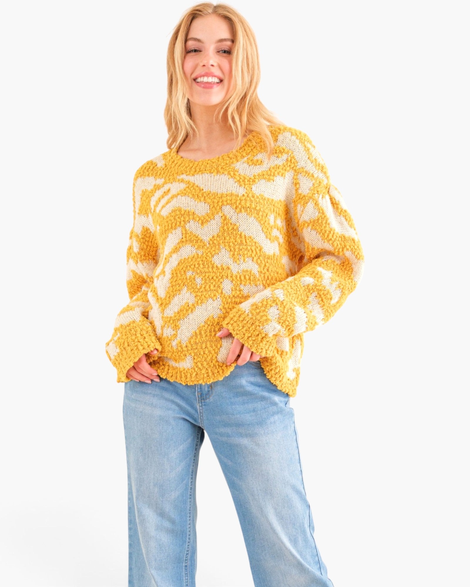 A woman wearing a yellow and cream wavy knit sweater with light blue jeans, standing against a plain white background.