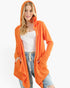 A woman modeling a bright orange open-front hooded cardigan over a white ribbed tank top and light blue jeans, photographed against a plain light gray studio background.