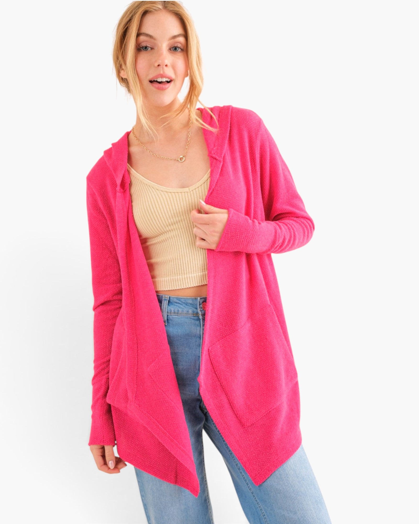 A woman modeling a bright pink open-front cardigan over a beige ribbed tank top and blue jeans, photographed against a plain light gray studio background.
