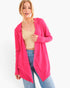 A woman modeling a bright pink open-front cardigan over a beige ribbed tank top and blue jeans, photographed against a plain light gray studio background.