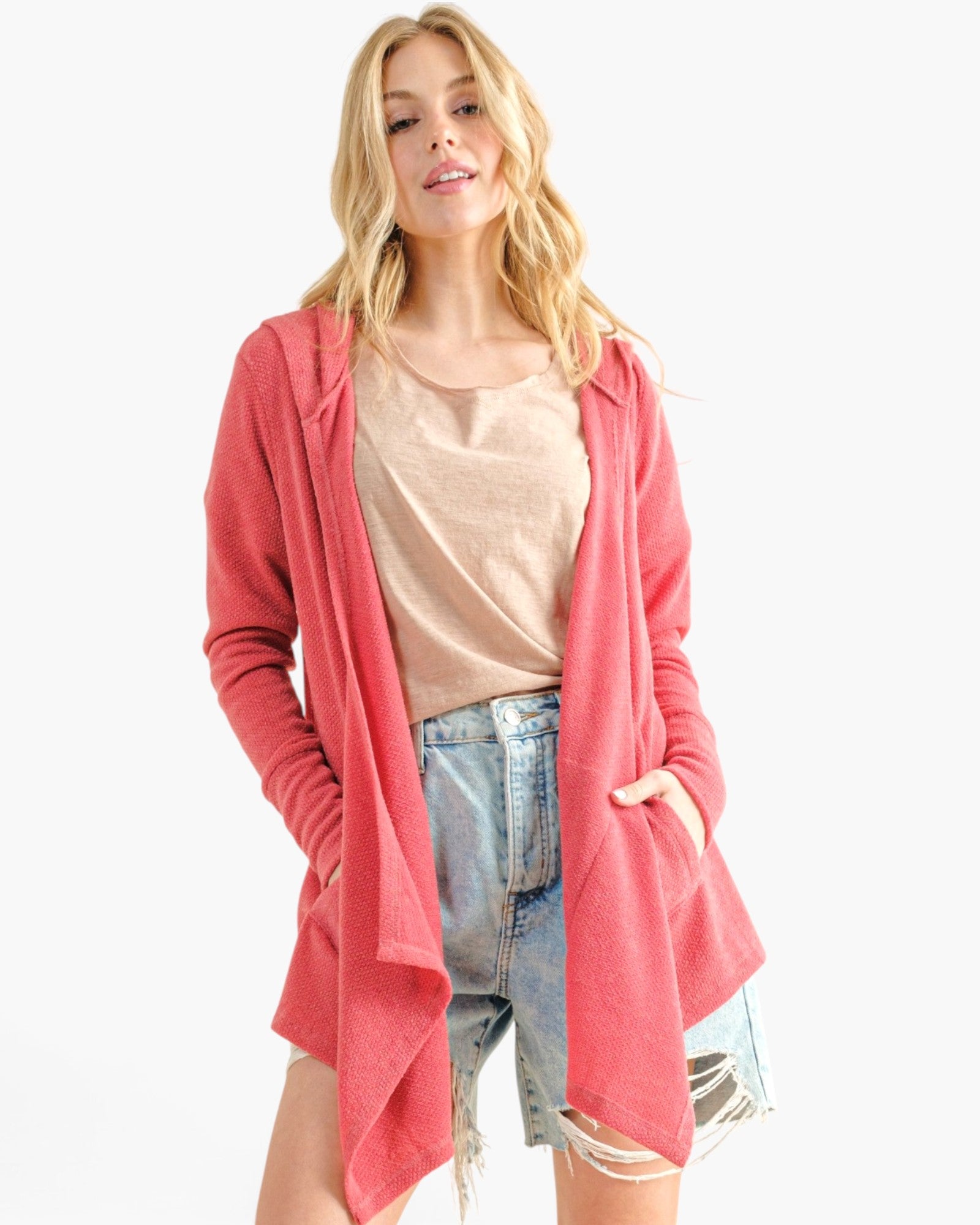 A woman modeling a coral pink open-front cardigan over a beige t-shirt and denim shorts, set against a plain white studio background.