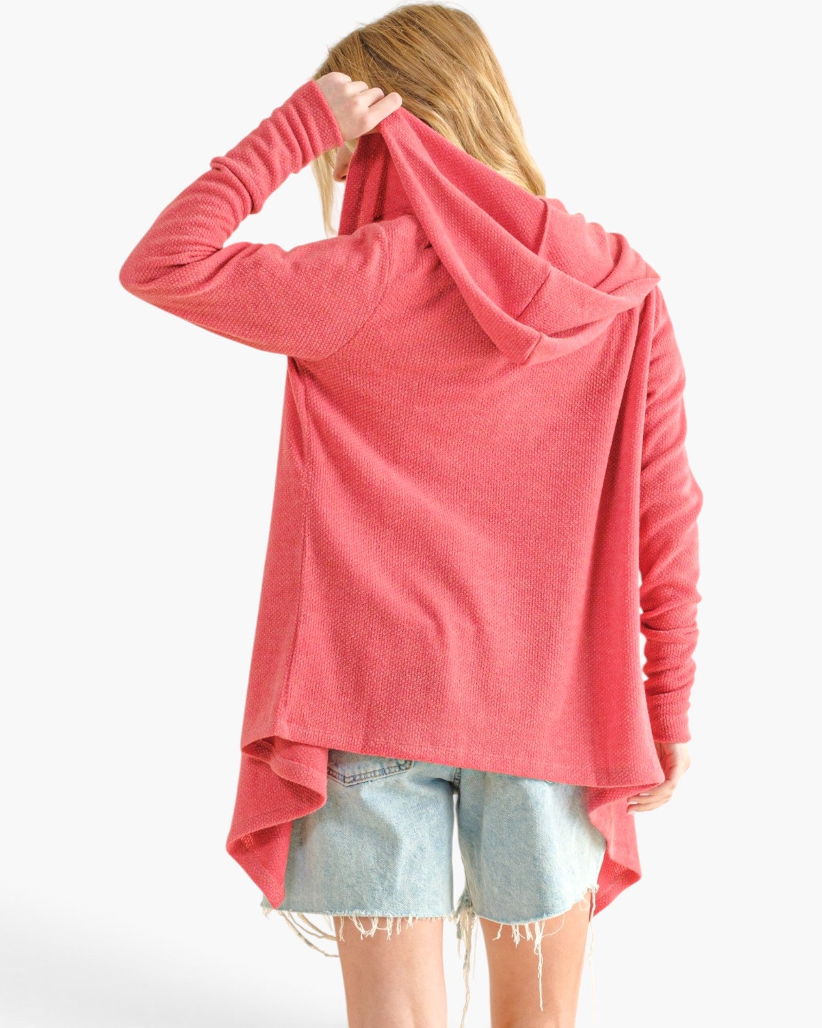 A woman shown from the back wearing a coral pink long-sleeve hooded top and light-wash denim shorts against a plain white studio background.