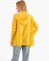 A woman shown from the back wearing a bright yellow hooded sweatshirt and light blue jeans against a plain white studio background.
