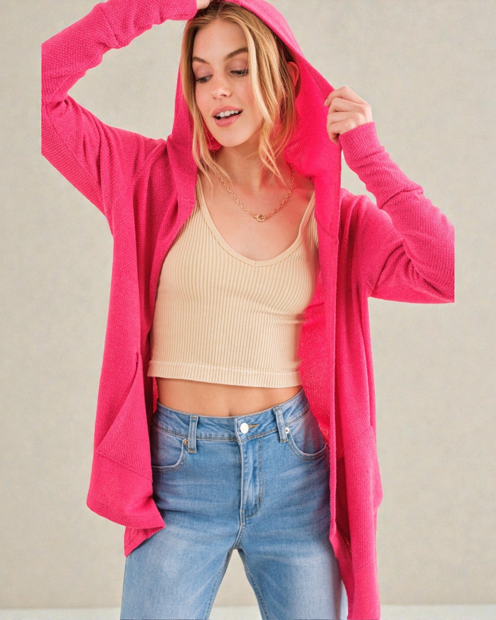 Woman wearing a bright pink hoodie, beige tank top, and blue jeans against a plain background