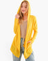 A woman wearing a bright yellow open-front hooded cardigan over a beige top and light blue jeans, photographed against a plain light gray studio background.