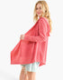 A woman wearing a coral pink open-front long-sleeve cardigan with a hood and large front patch pockets, paired with light denim shorts, photographed against a plain white studio background.