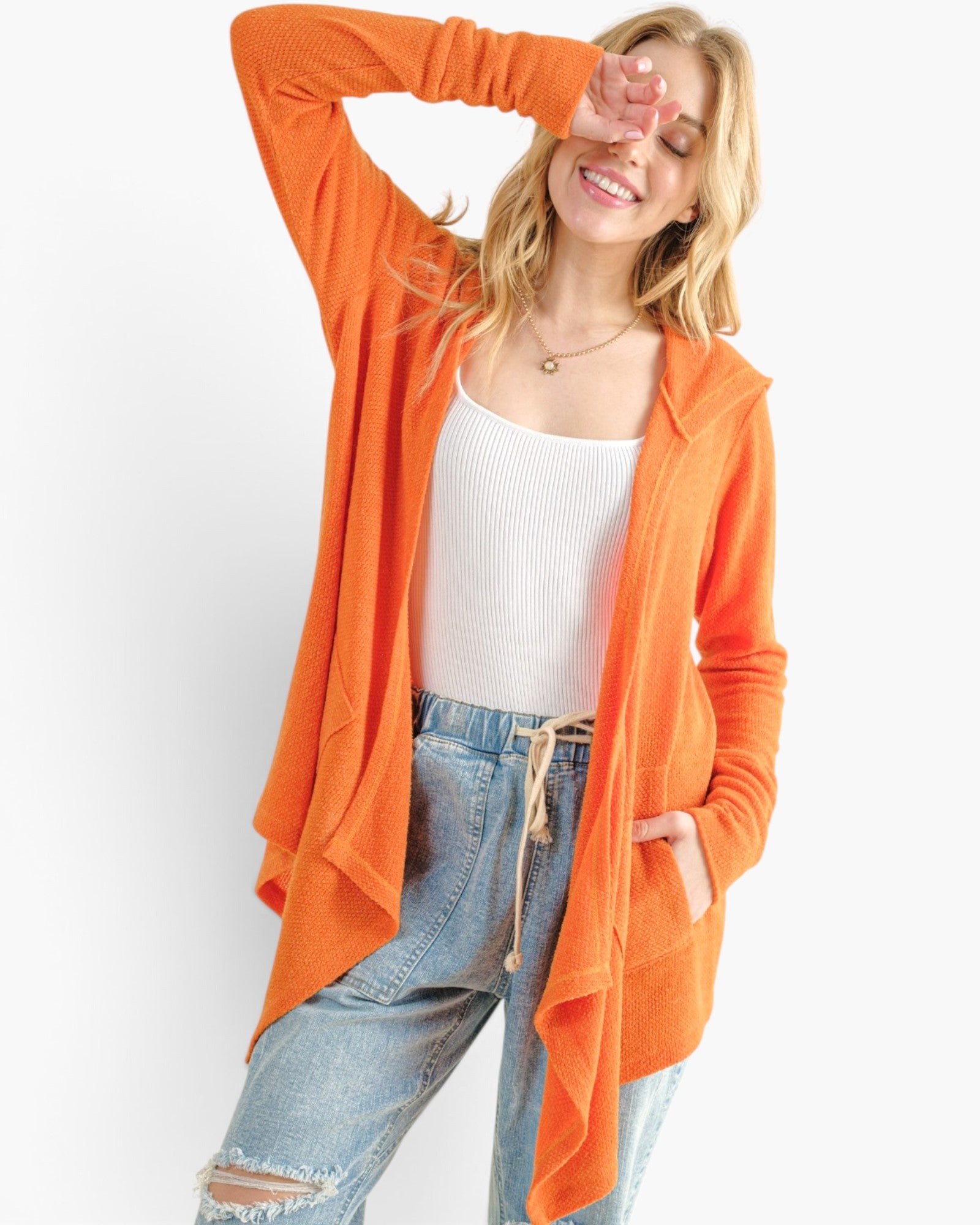 A woman modeling a bright orange open-front cardigan over a white ribbed tank top and light blue jeans, posed against a clean white studio background.
