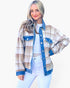 A woman modeling a long-sleeve plaid overshirt with blue denim trim, worn over a white top and white pants against a light gray studio background.