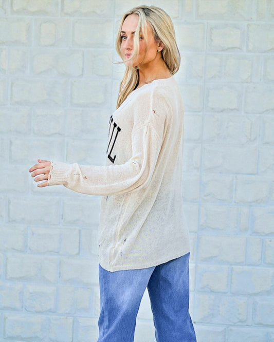 Woman wearing a cream sweater with text and blue pants against a light brick wall.