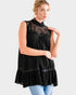 A woman modeling a sleeveless black tunic top with a lace ruffled mock neck and tiered ruffle hem, paired with light blue jeans against a light gray studio background.