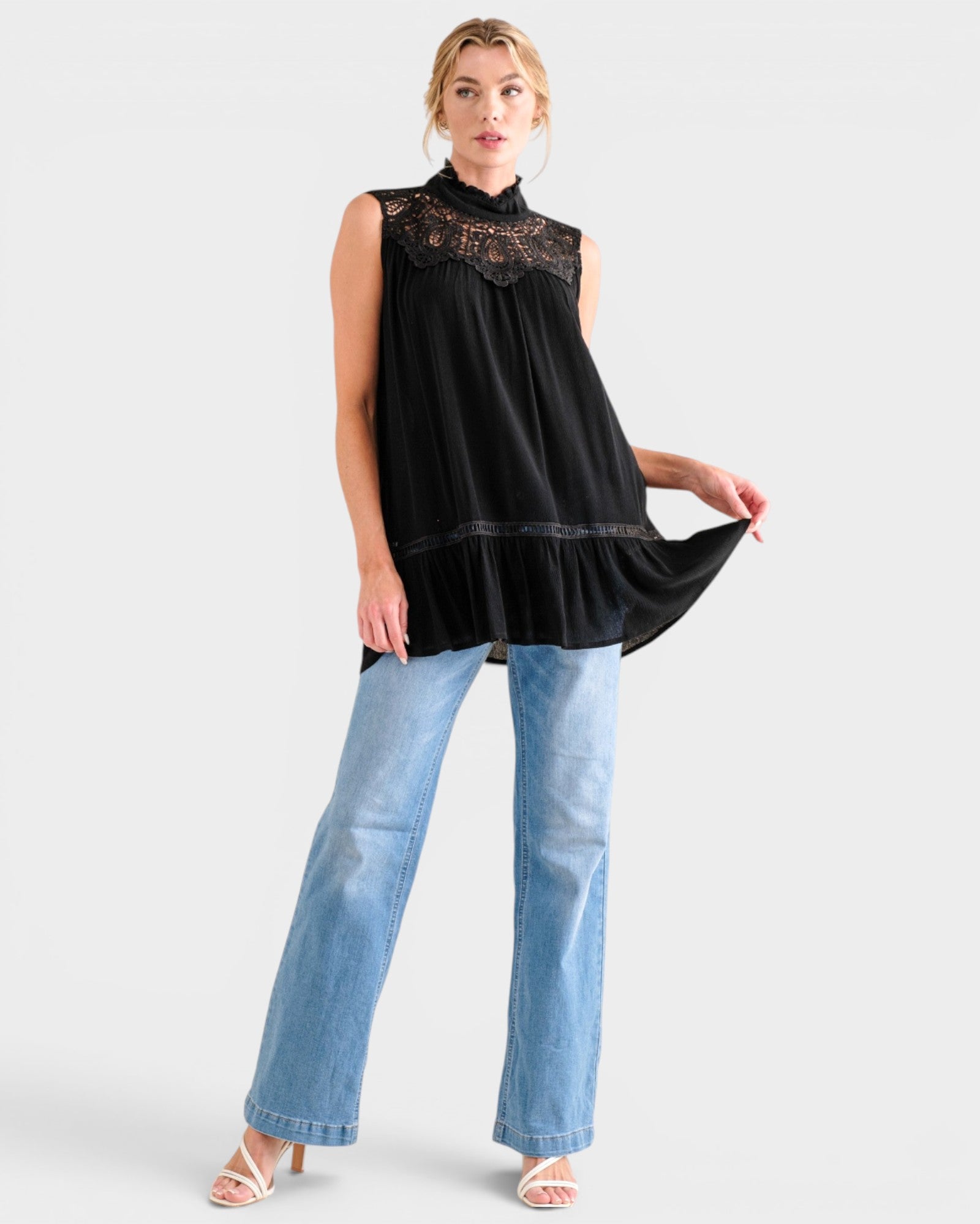 A woman modeling a sleeveless black top with a lace high neckline and tiered skirt, paired with light blue wide-leg jeans and white heeled sandals, against a light gray studio background.