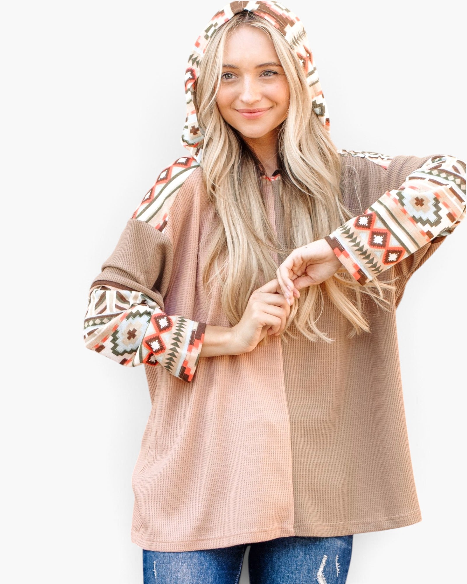 A woman modeling a colorblock hooded pullover top with a solid taupe-brown body, a dusty pink lower body, and patterned sleeves and hood in beige, brown, and red geometric designs, paired with blue jeans against a white background.