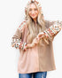 A woman modeling a colorblock hooded pullover top with a solid taupe-brown body, a dusty pink lower body, and patterned sleeves and hood in beige, brown, and red geometric designs, paired with blue jeans against a white background.