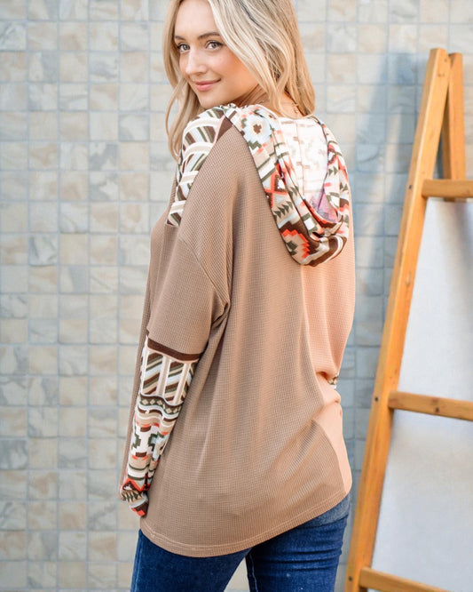 Woman wearing a beige sweater with patterned sleeves against a tiled wall.