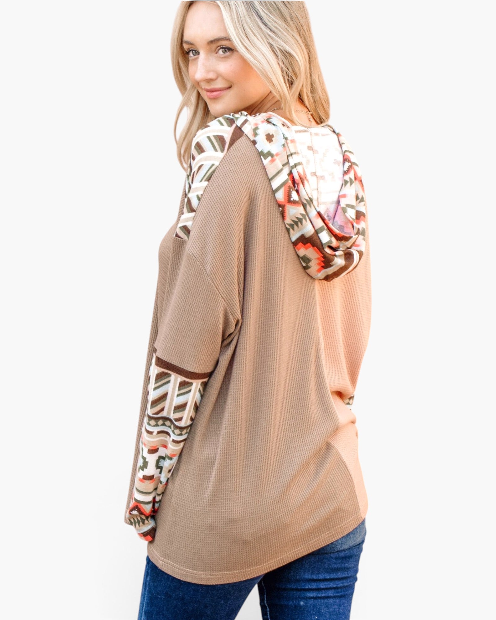 A woman wearing a waffle-knit long-sleeve top with a solid tan body and a colorful geometric patterned hood, paired with blue jeans against a light gray background.