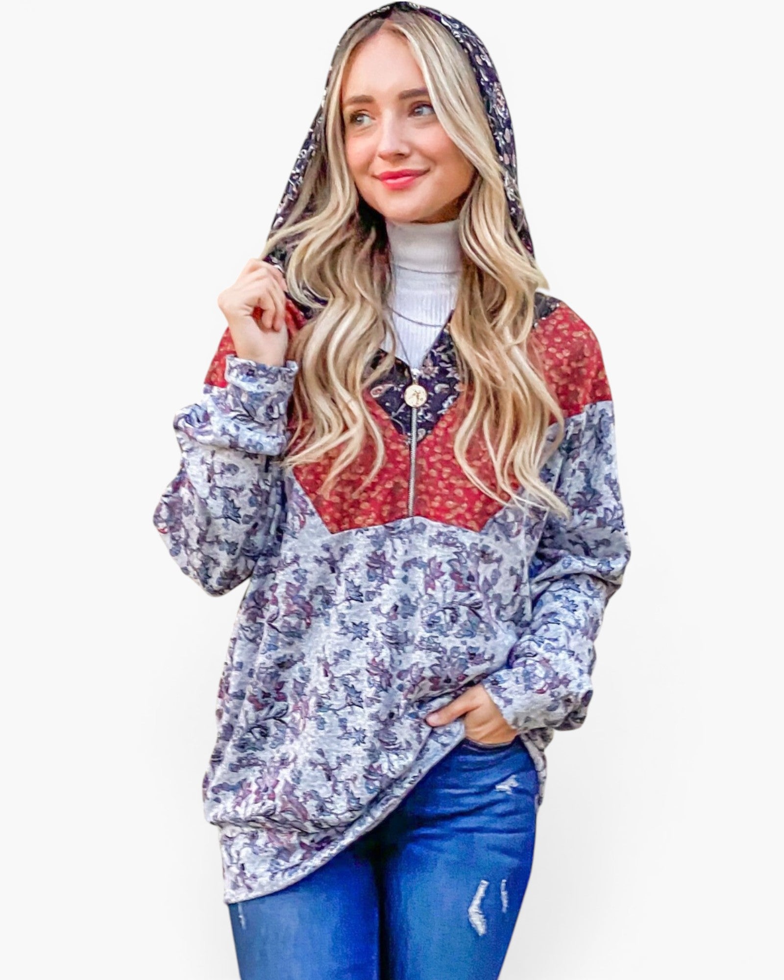 A woman wearing a hooded zip-up jacket with a blue and purple floral pattern, red color-blocked yoke, and a white turtleneck underneath, paired with blue jeans, against a plain light gray background.