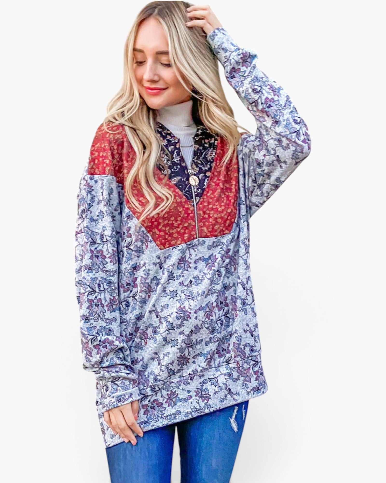 A woman modeling a long-sleeve half-zip pullover jacket with a blue and purple floral pattern, red patterned yoke panels, and a dark patterned collar, paired with blue jeans against a light gray background.
