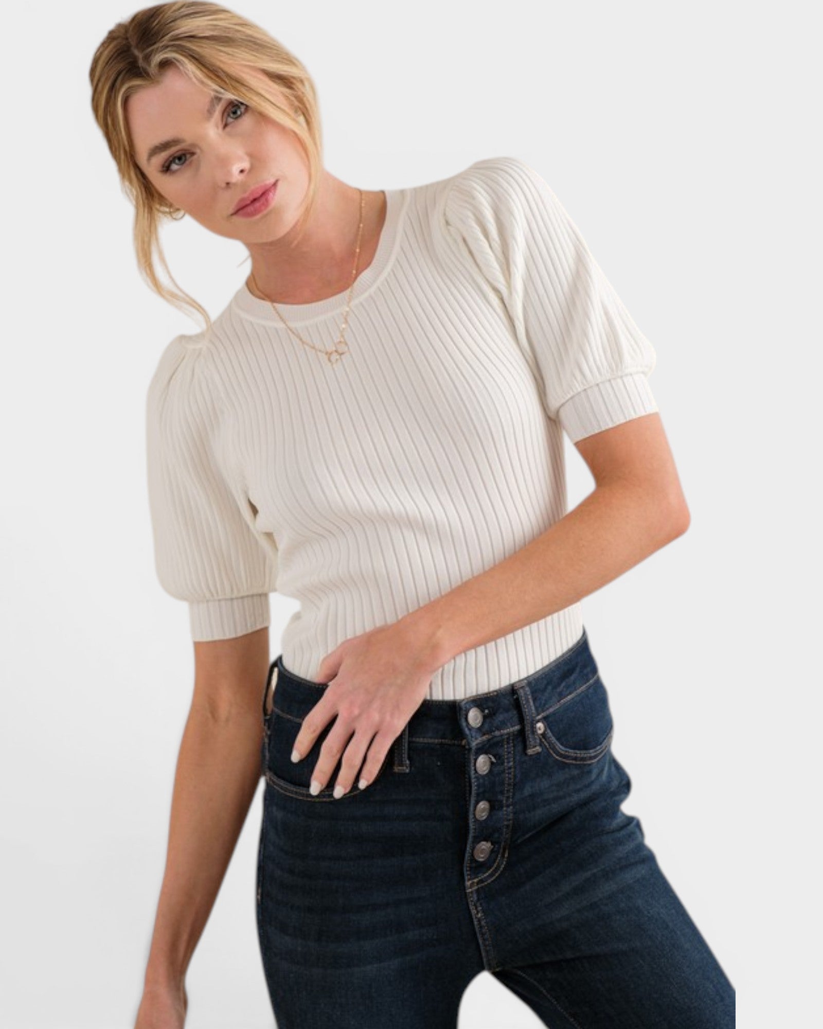 Woman wearing a white ribbed sweater and dark jeans on a plain background