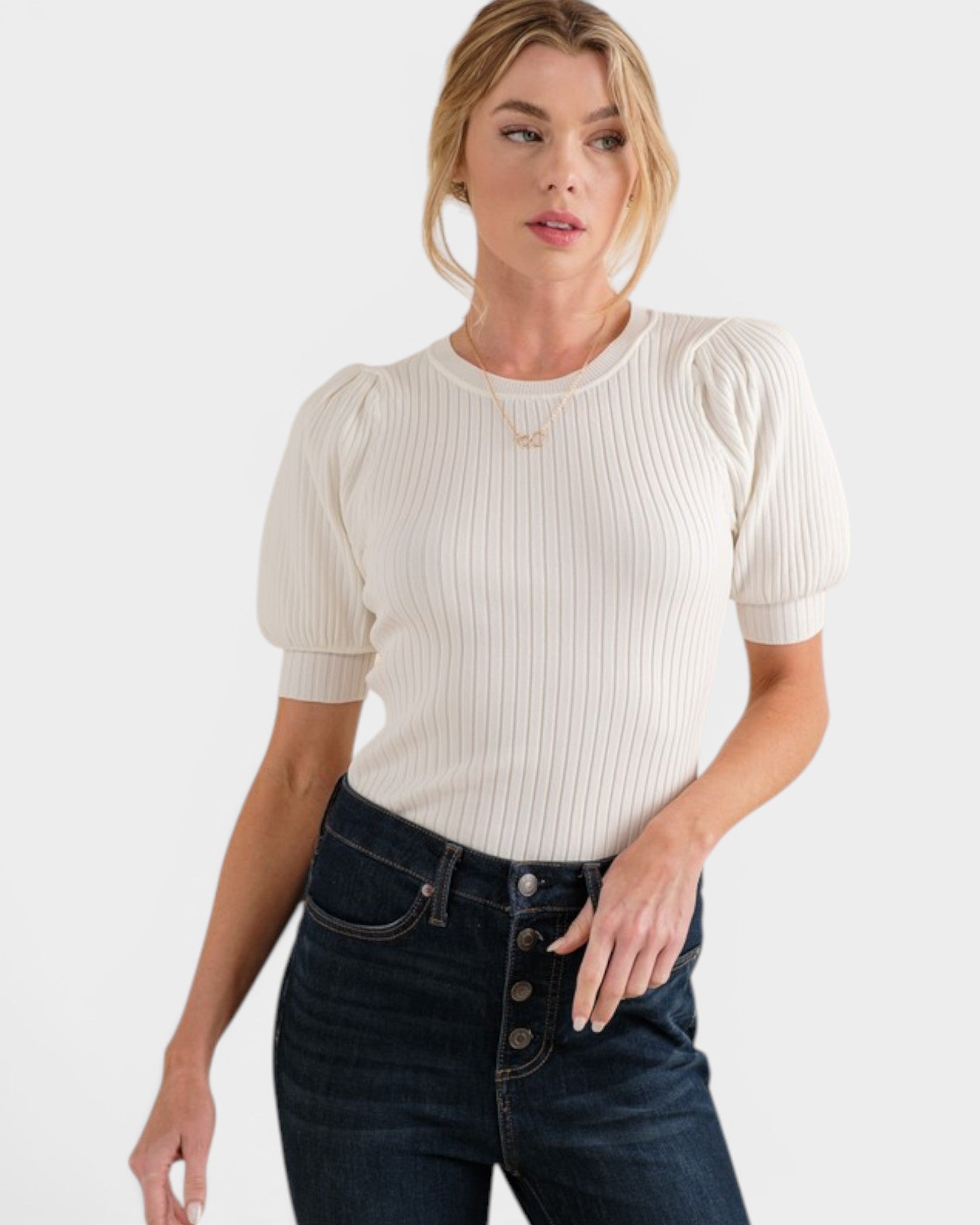 Woman wearing a cream ribbed top with puff sleeves and dark jeans on a white background