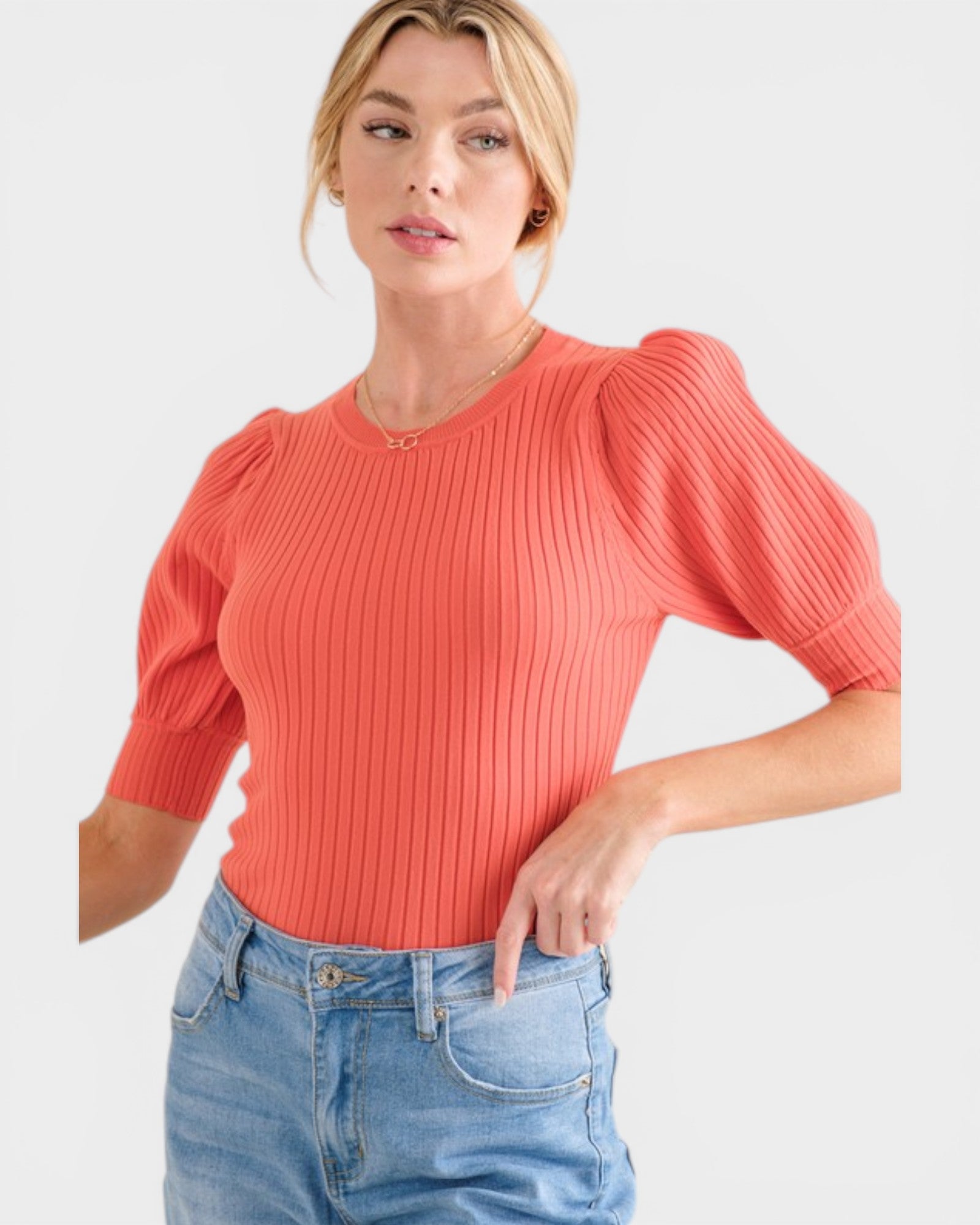 Woman wearing a coral ribbed top with puff sleeves and blue jeans on a light gray background