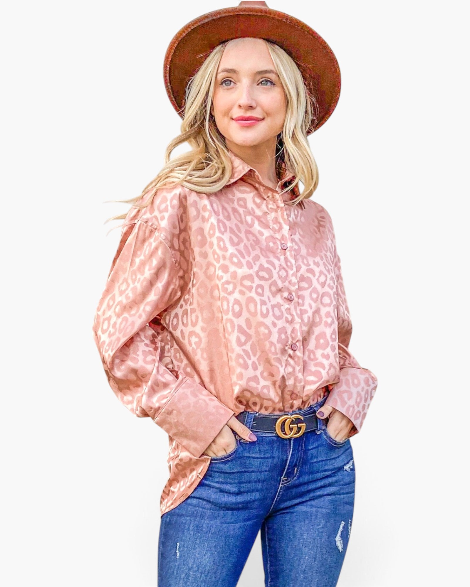 A woman modeling a pink leopard-print satin button-up shirt, styled with a brown wide-brim hat, a black belt with a gold buckle, and blue jeans, against a plain white background.