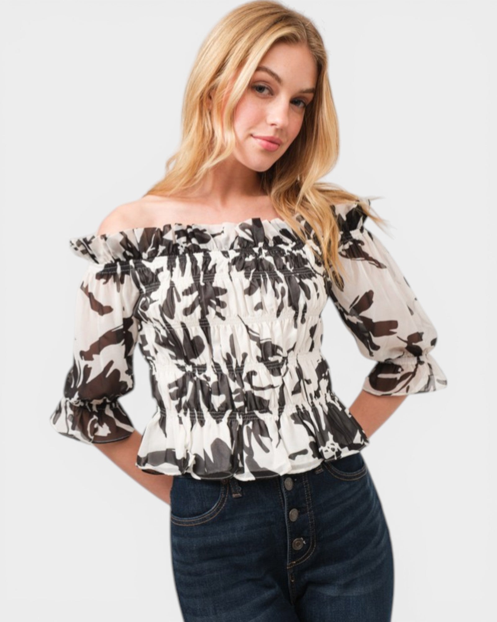 Woman wearing a black and white floral off-shoulder top with blue jeans on a light gray background