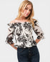 Woman wearing a black and white floral off-shoulder top with blue jeans on a light gray background