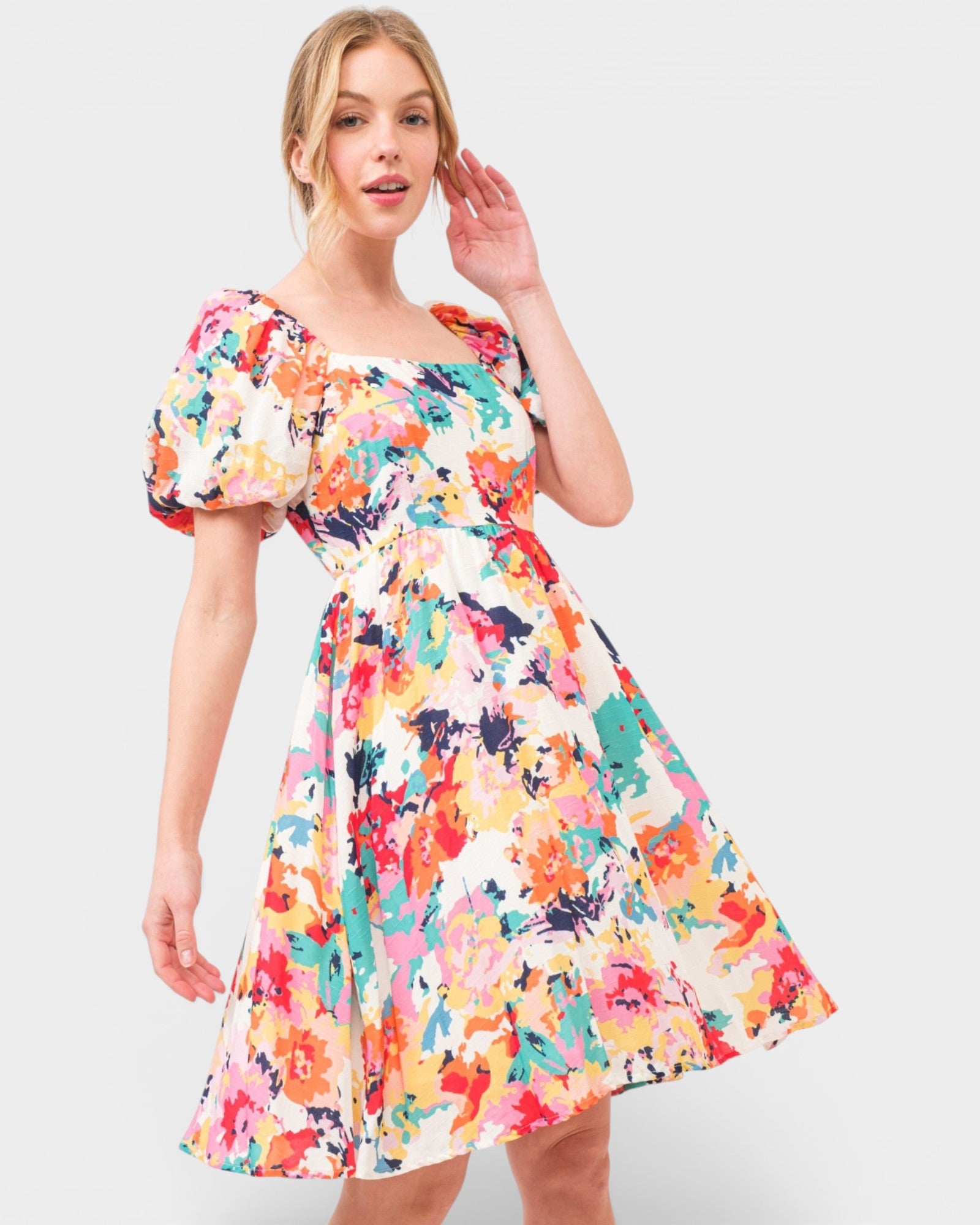 A woman modeling a short puff-sleeve A-line dress with a vibrant multicolor floral and abstract watercolor print on a cream background, set against a plain light gray studio background.