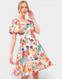 A woman modeling a short puff-sleeve A-line dress with a vibrant multicolor floral and abstract watercolor print on a cream background, set against a plain light gray studio background.