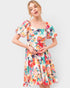 A woman modeling a short puff-sleeve dress with a square neckline and a vibrant multicolor floral print on a light cream background, set against a plain light gray studio backdrop.