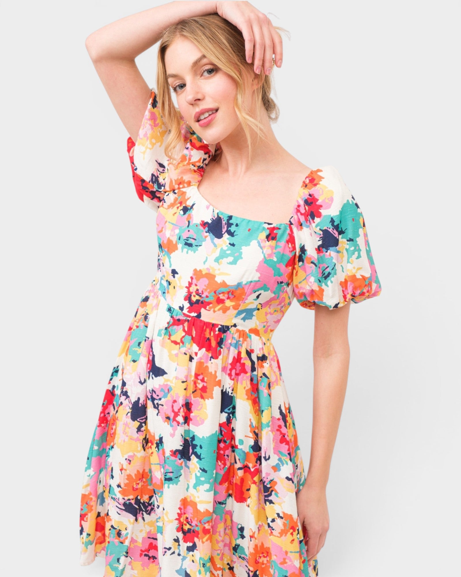 A woman modeling a short puff-sleeve dress with a colorful abstract floral print on a light background, set against a plain light gray studio backdrop.